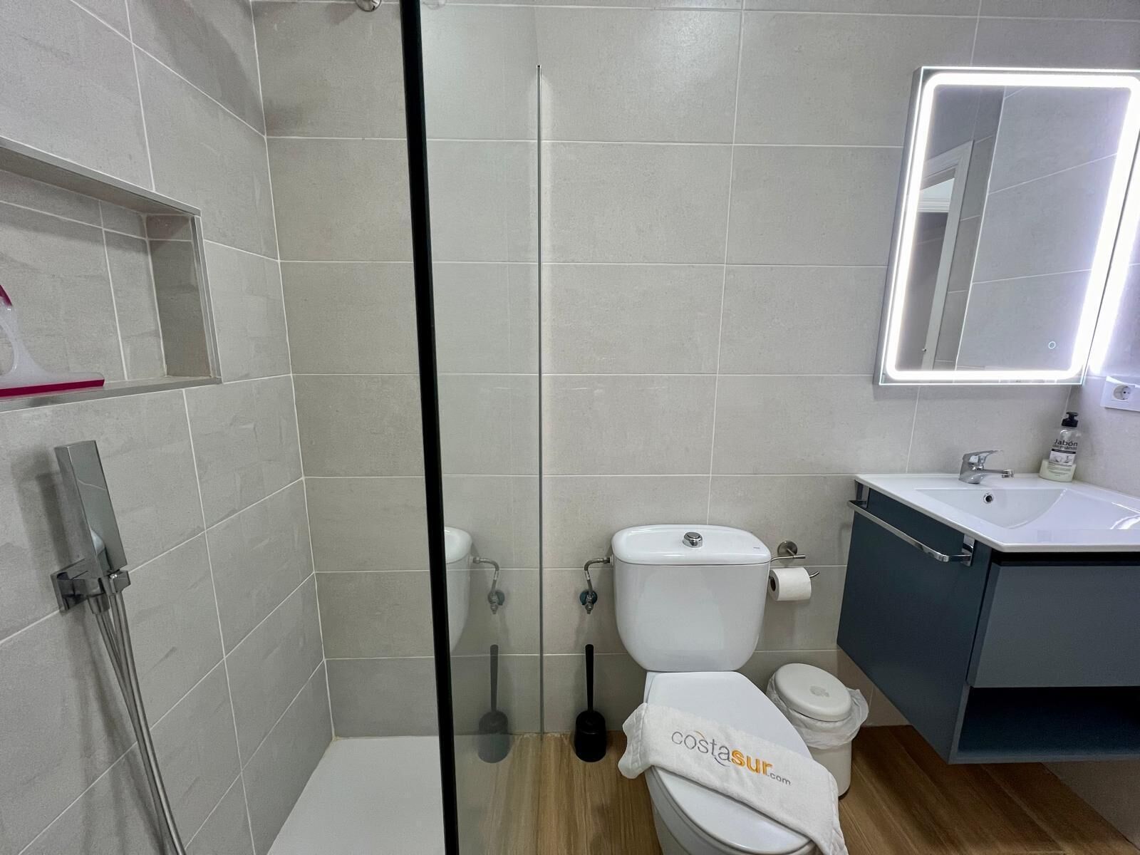 A modern bathroom with a glass-enclosed shower, a white toilet, a sink with a mirror, and a shelf with toiletries.