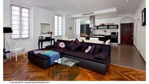 Ein modernes Wohnzimmer mit einer dunklen Couch, einem Glastisch und einem Essbereich mit weißem Tisch und Stühlen.