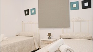 A hotel room with two beds, each with a white bedspread and matching pillows. There is a nightstand with a lamp and a folded towel on it. The walls have two framed pictures, one black and one teal. - Rural apartment for four people (CONIL DE LA FRONTERA)