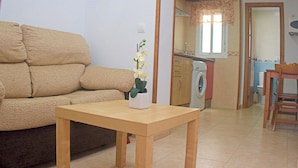 A living room with a beige sofa, a wooden table with a vase of flowers, a washing machine, and a dining area with a chair. - Central apartment with air conditioning (CONIL DE LA FRONTERA)