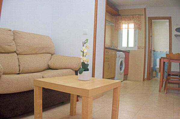Ein Wohnzimmer mit einem beigen Sofa, einem Holztisch mit Blumenvase, einer Waschmaschine und einem Essbereich mit einem Stuhl.