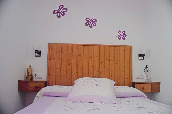 Ein Schlafzimmer mit Holz-Kopfteil, einem Bett mit floralem Bettbezug, zwei Nachttischen mit Lampen und blumengeschmückter Wanddeko.