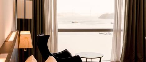A hotel room with a bed, a chair, a small table, and a view of a body of water through large windows.