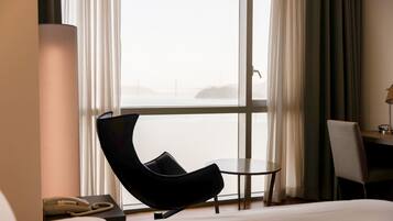 A hotel room with a bed, a chair, a small table, a lamp, and a phone.