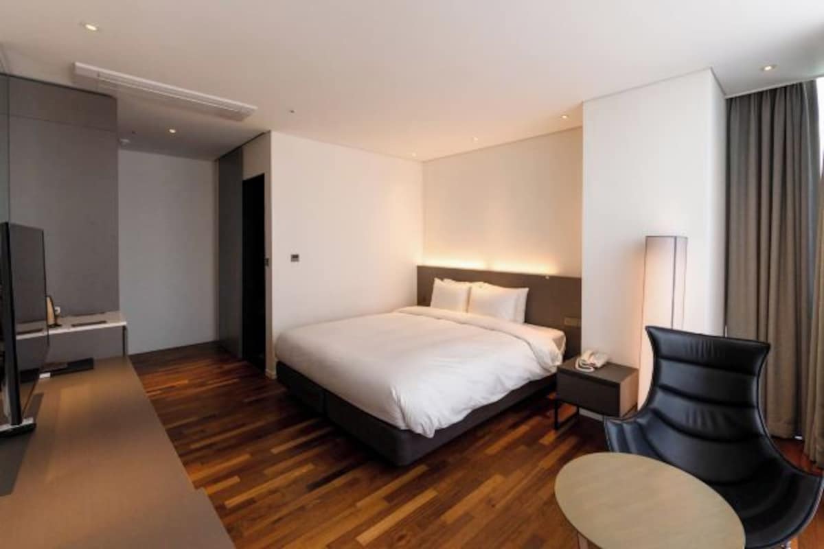 Una habitación de hotel moderna con una cama grande, una mesita de noche con lámpara, una silla negra y un televisor sobre un mueble.