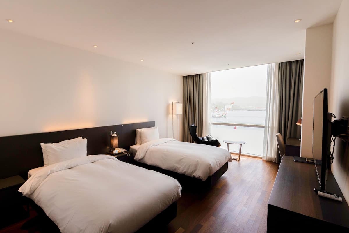 Ett hotellrum med två sängar, ett skrivbord, en stol, en TV och utsikt över staden genom stora fönster.