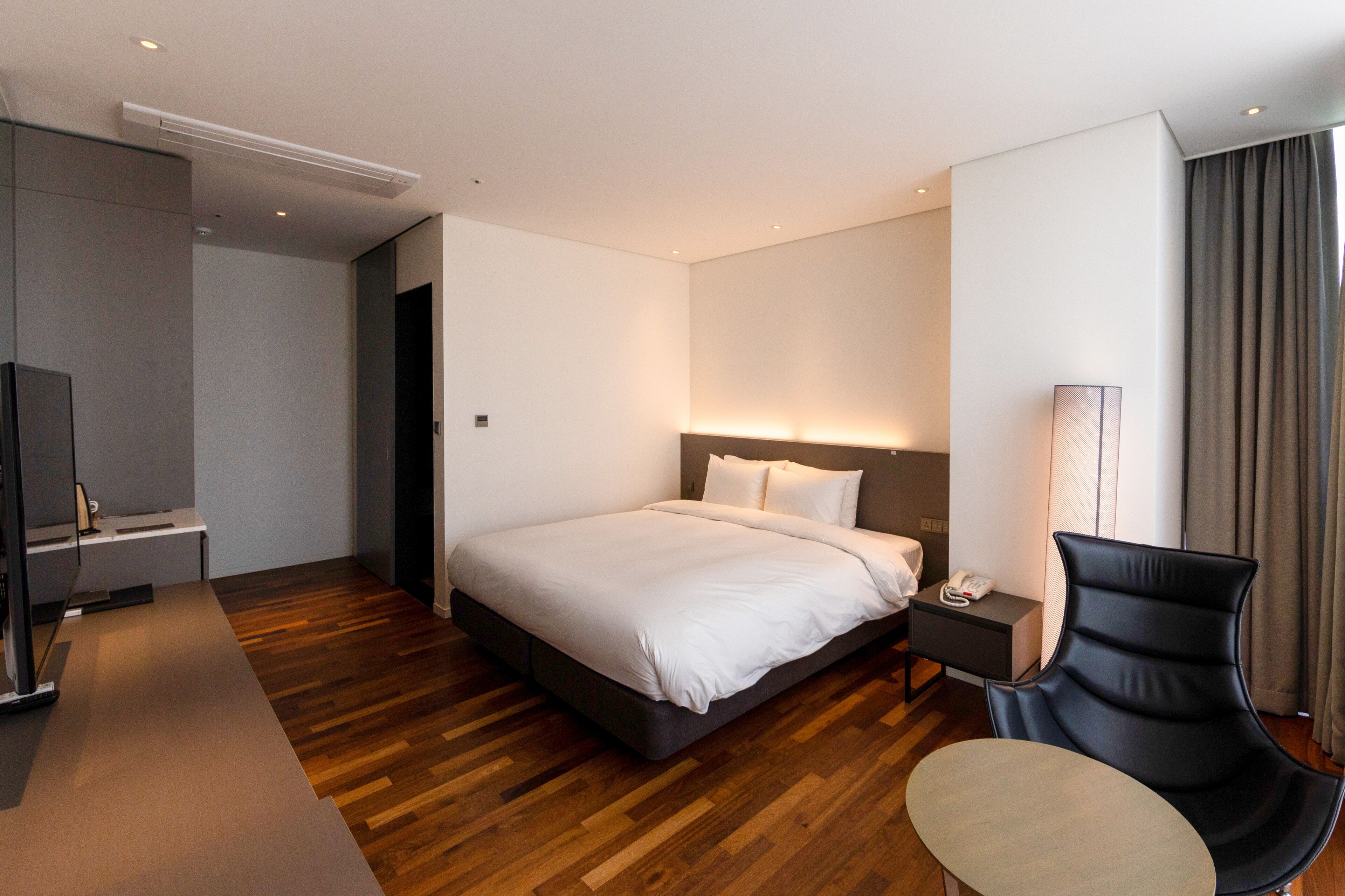 A modern hotel room with a large bed, bedside table, chair, and a desk with a lamp.