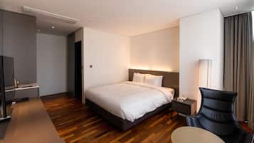A modern hotel room with a large bed, bedside table, chair, and a desk with a lamp.
