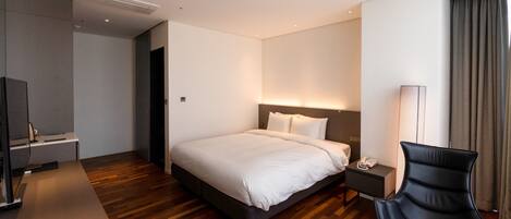 A modern hotel room with a large bed, bedside table, chair, and a desk with a lamp.