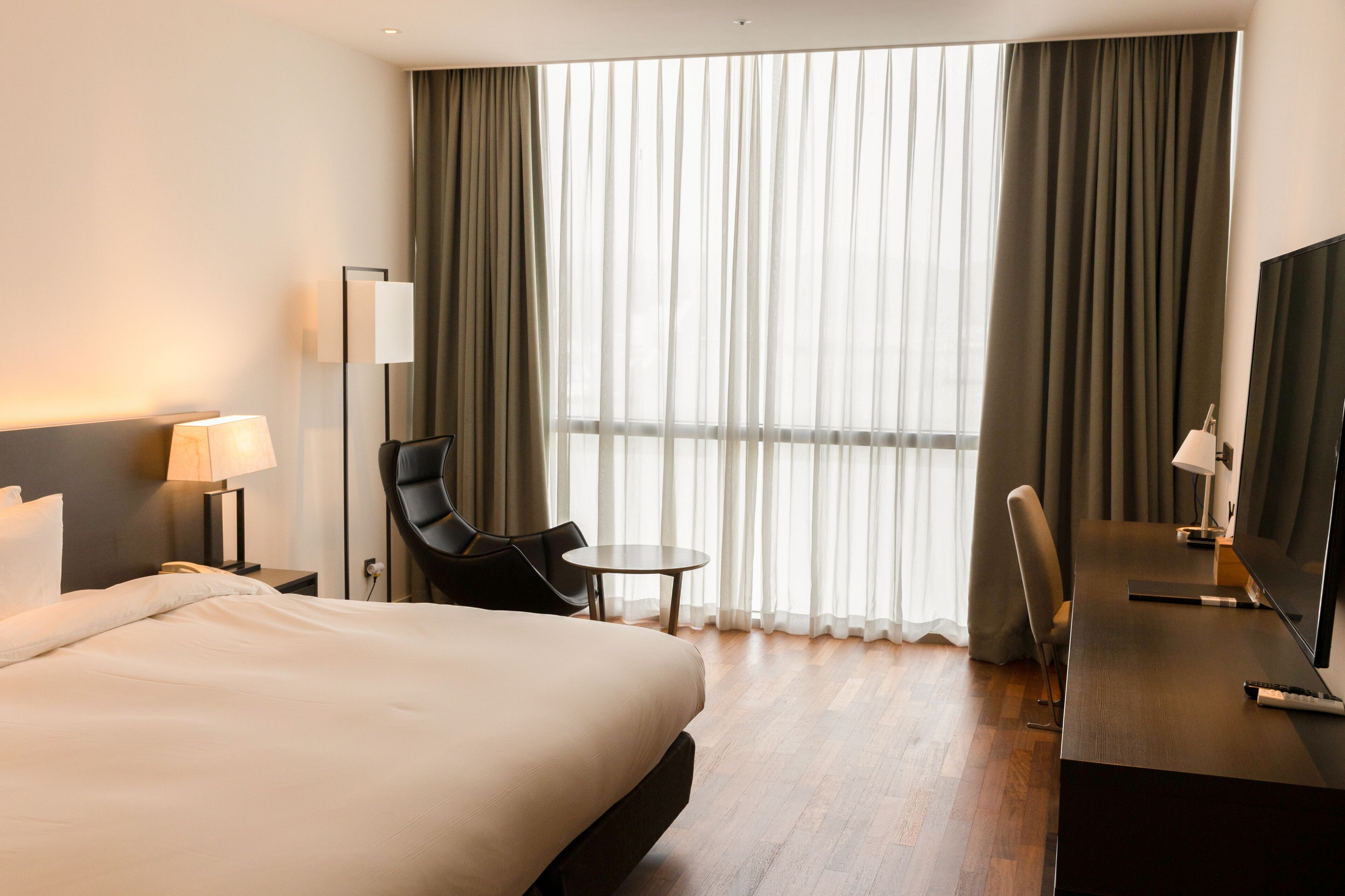 A hotel room with a bed, a desk, a chair, a TV, and a window with curtains.
