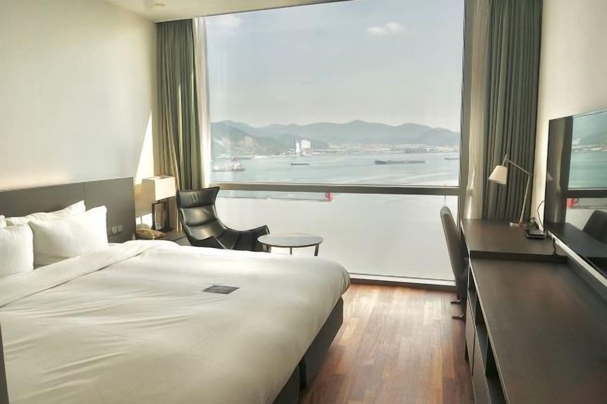 Habitación de hotel con una cama grande, un escritorio, una silla y vistas al agua y a las montañas.