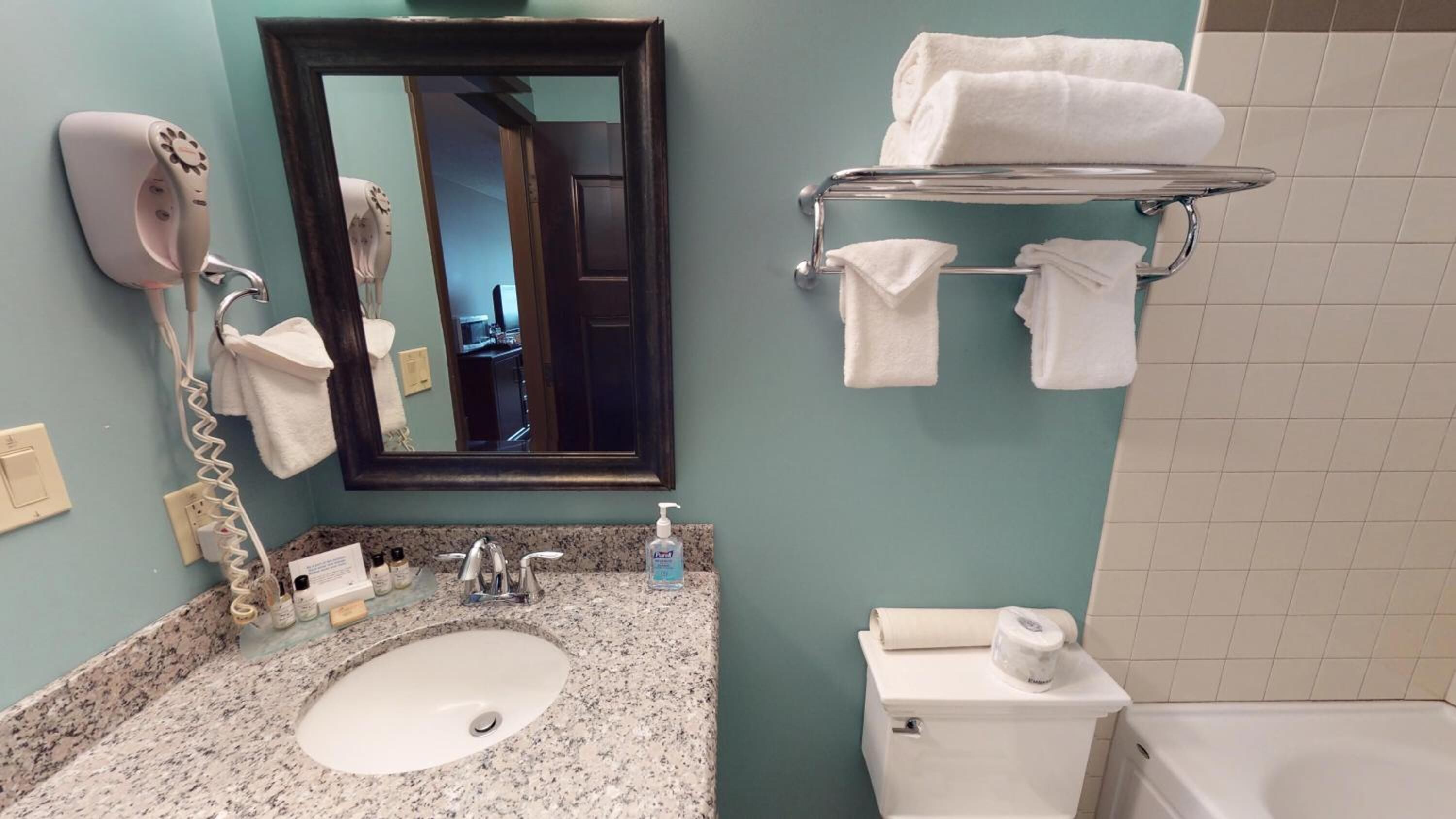 A bathroom with a granite countertop sink, a mirror, a hairdryer, a towel rack, and a toilet.