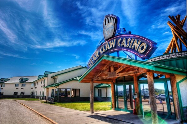 A casino entrance with a claw machine sign, a covered waiting area, and a building with multiple windows in the background. - Bear Claw Casino & Hotel (Carlyle)