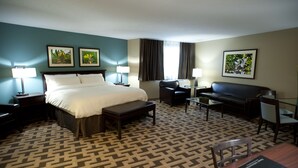 Habitación de hotel con una cama grande, un escritorio, un sofá y dos sillones. Hay dos lámparas en las mesitas de noche y una ventana con cortinas.