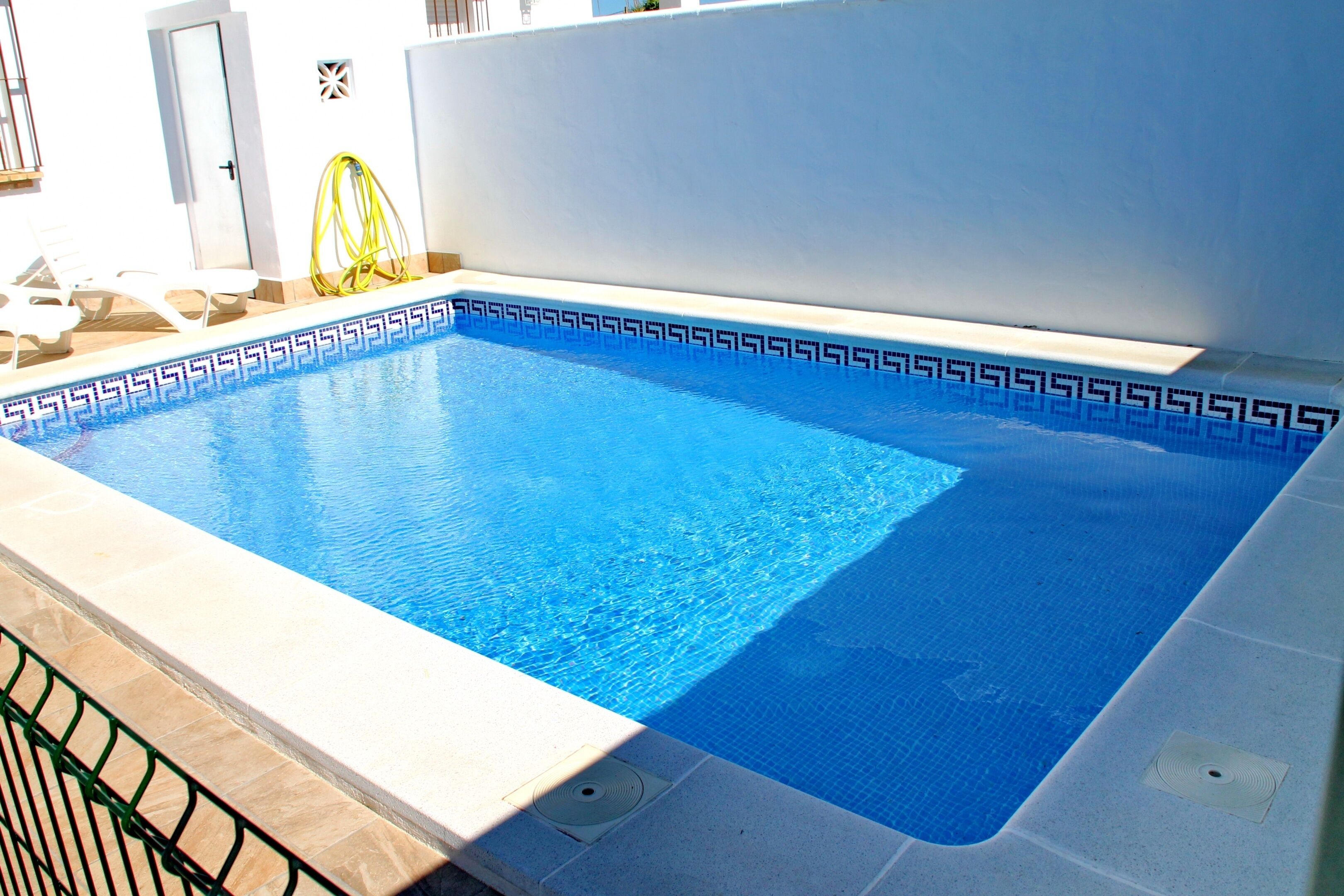 Une piscine rectangulaire bordée de carreaux aux motifs géométriques, un tuyau jaune et une porte visible au fond.