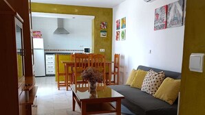 Un salon avec un canapé, une table à manger entourée de chaises, une cuisine en arrière-plan et un mur jaune.