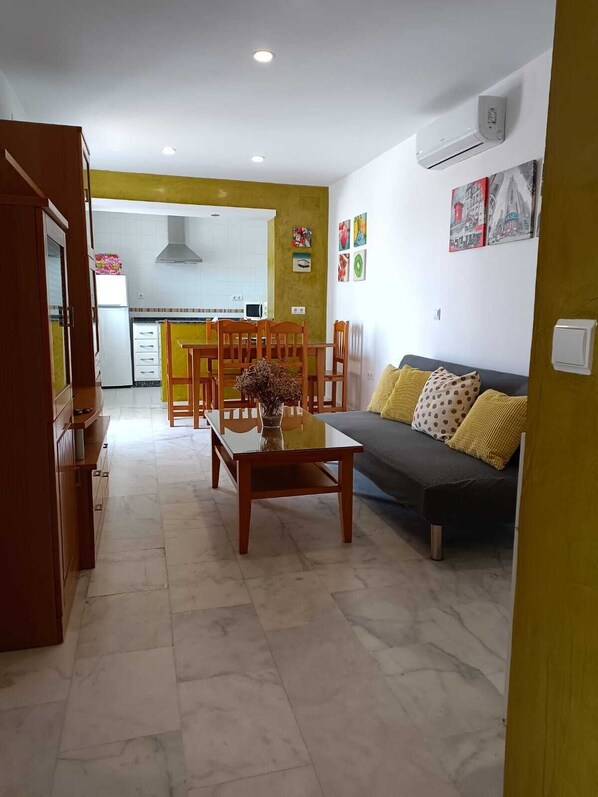 A living room with a sofa, a dining table with chairs, a kitchen in the background, and a yellow wall.