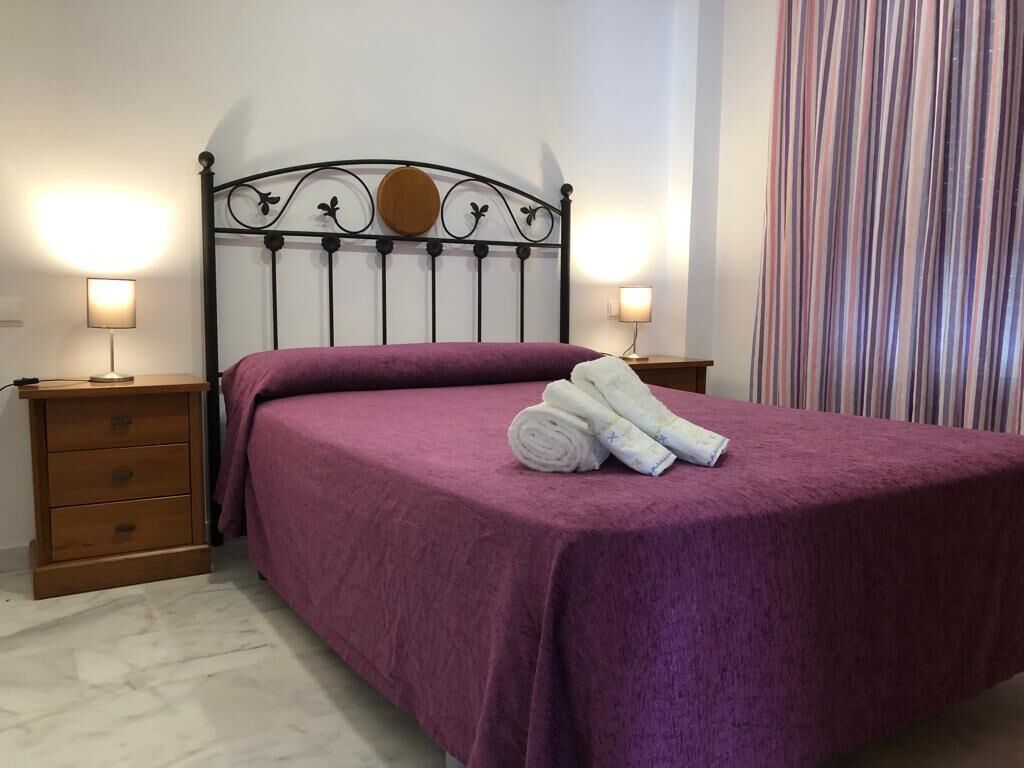 A neatly made bed with a purple bedspread, two bedside tables with lamps, and a striped curtain.