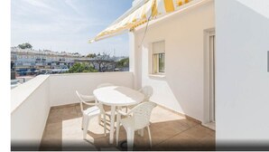 Un balcon avec une table et des chaises blanches, un auvent à rayures jaunes et une vue sur des bâtiments au loin.