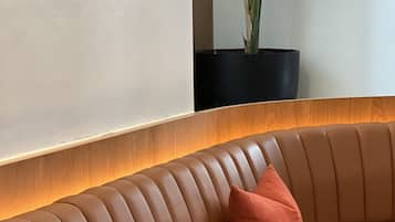 Un sofá de cuero moderno y curvo con dos cojines, una mesita de noche negra pequeña y una planta en maceta frente a una pared blanca.