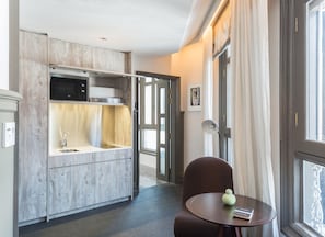 A compact kitchen with a microwave, sink, and wooden cabinets. There is a dining area with a table and a chair, and a window with curtains.