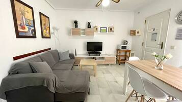 Een woonkamer met een bank, televisie, salontafel en een eethoek met stoelen.