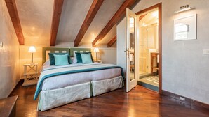 A bedroom with a large bed, wooden beams on the ceiling, a walk-in closet, and a bathroom visible through an open door.