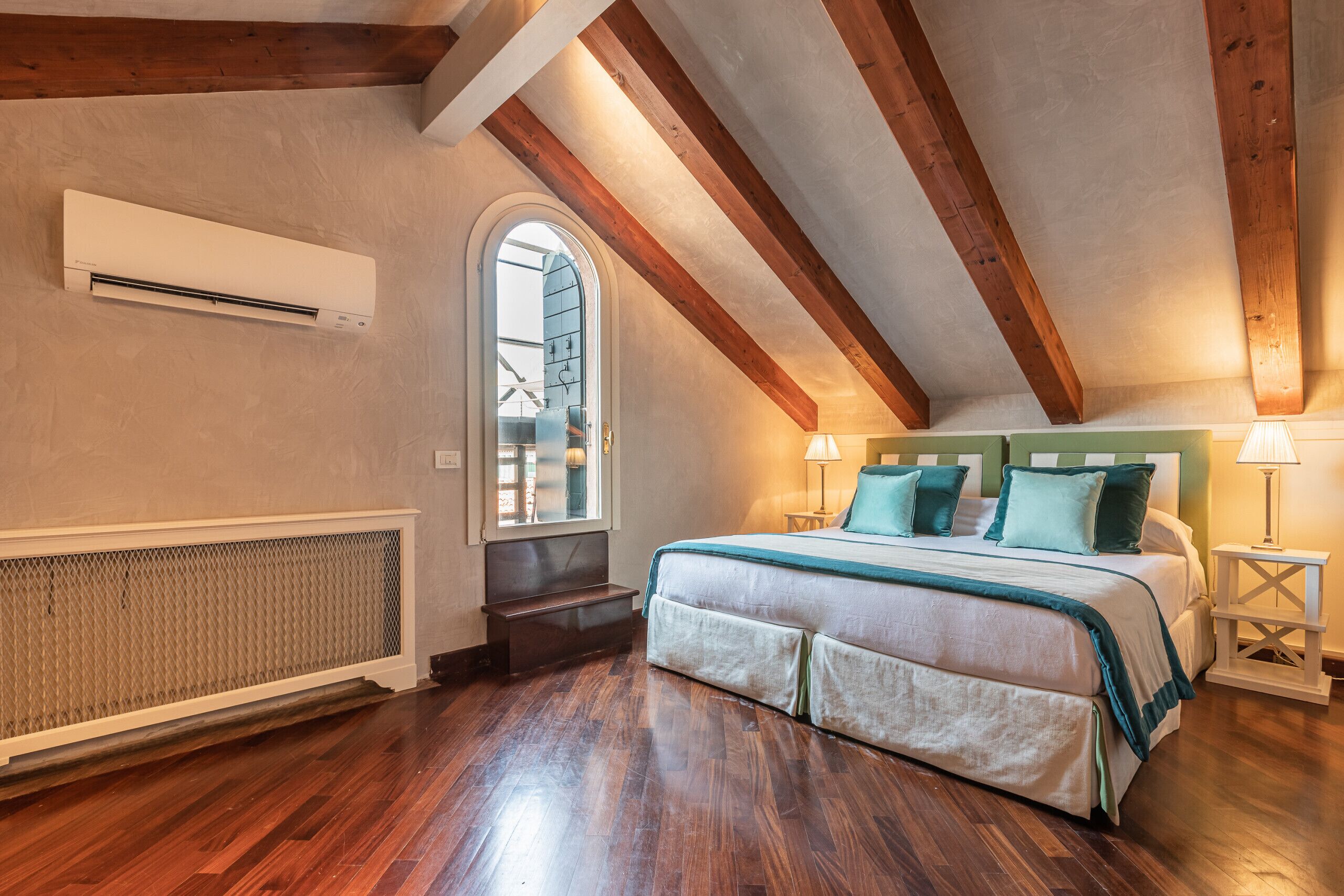 Een slaapkamer met een houten plafond, een groot bed met groen en wit beddengoed, een raam en een radiator.