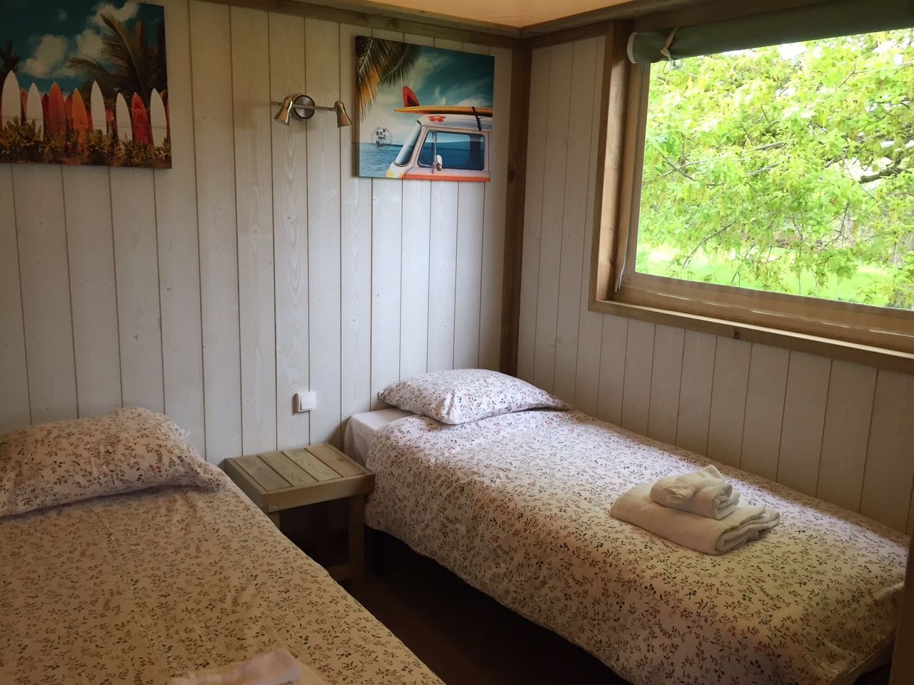 A small, cozy room with two single beds, a wooden nightstand, and a window with a view of greenery.