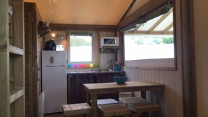 A compact kitchen with wooden cabinets, a white refrigerator, a microwave, and a window with a view of greenery. There is a wooden table with a blue bowl and a small stool.