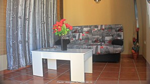 A living room with a sofa, a small table with a vase of flowers, a curtain with a floral pattern, and a wall-mounted air conditioner.