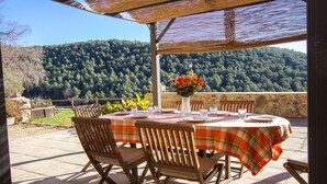 Une terrasse extérieure avec une table dressée pour le repas, entourée de chaises en bois, et offrant une vue sur une colline boisée.