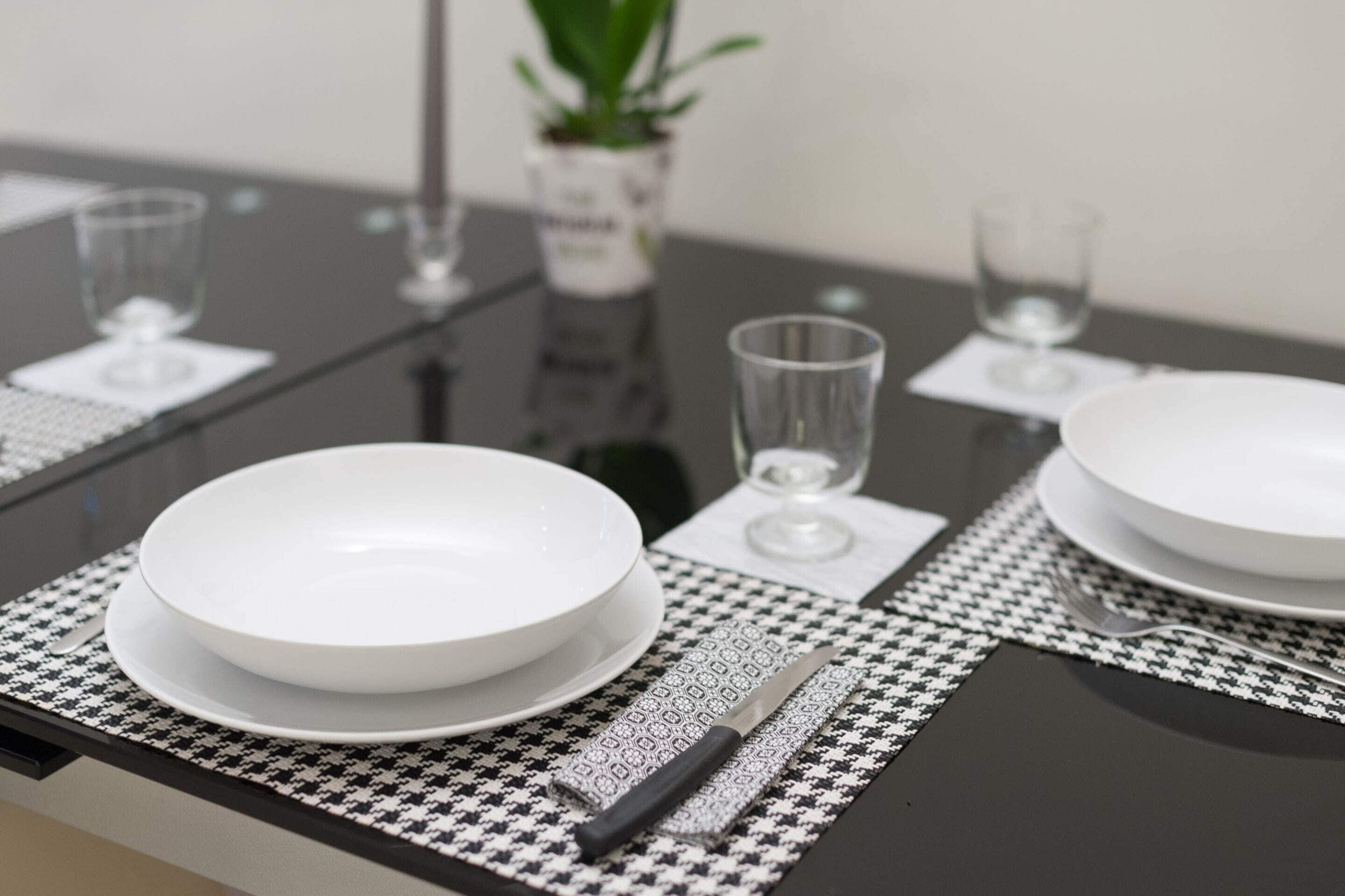 Une table à manger dressée avec de l’assiette blanche, des couverts et des verres, agrémentée d’un set de table à motifs noirs et blancs.