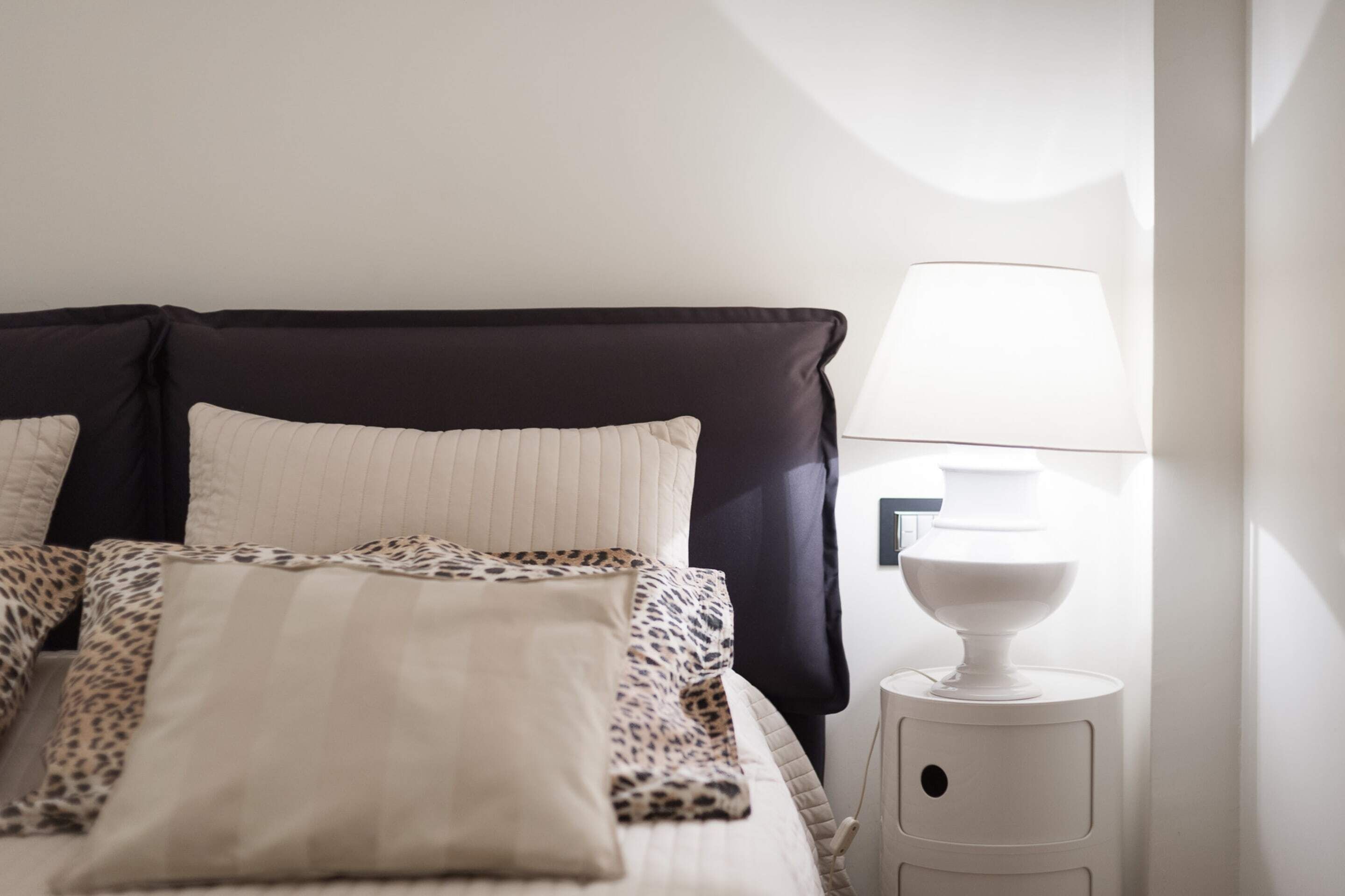 Een net opgemaakt bed met een donker hoofdbord, een witte nachtlamp en een kussen met luipaardprint.
