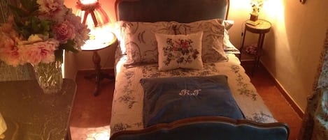 A bedroom with a bed, floral bedding, a vase of flowers, a lamp, and a mirror on the wall.