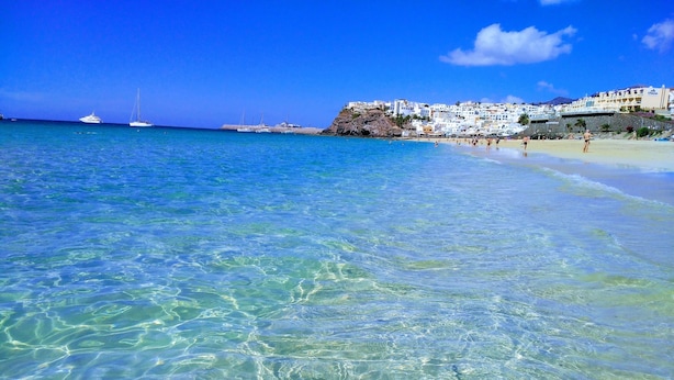 Una spiaggia con acque turchesi trasparenti, un edificio bianco con balconi e alcune imbarcazioni in lontananza.