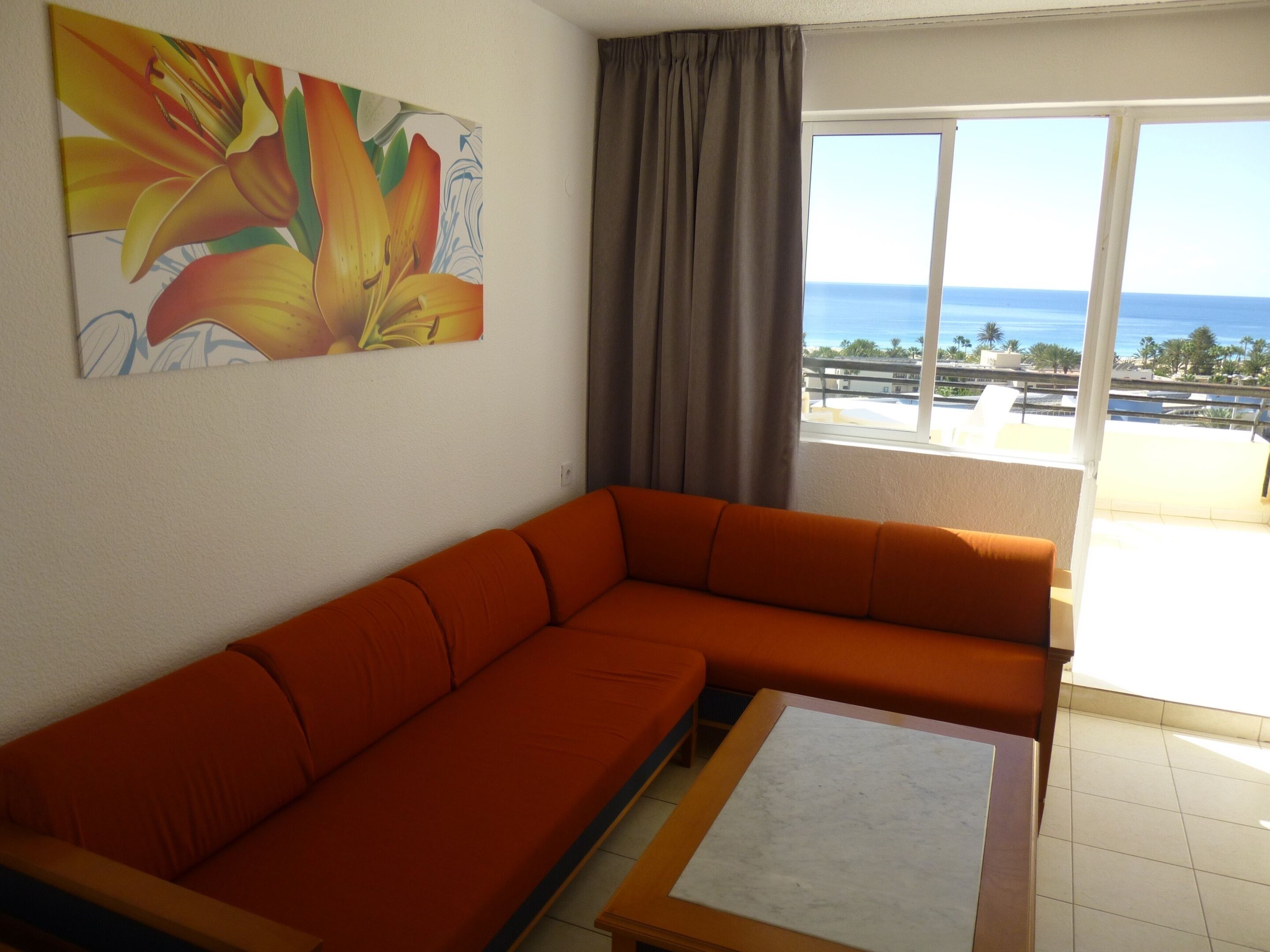 A living room with a sofa, a coffee table, and a painting of flowers. There is a window with a view of the ocean and buildings.