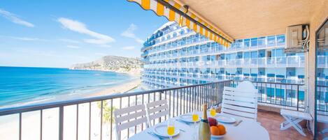 Ein Balkon mit Tisch für Zwei mit Blick auf Strand und Kreuzfahrtschiff.