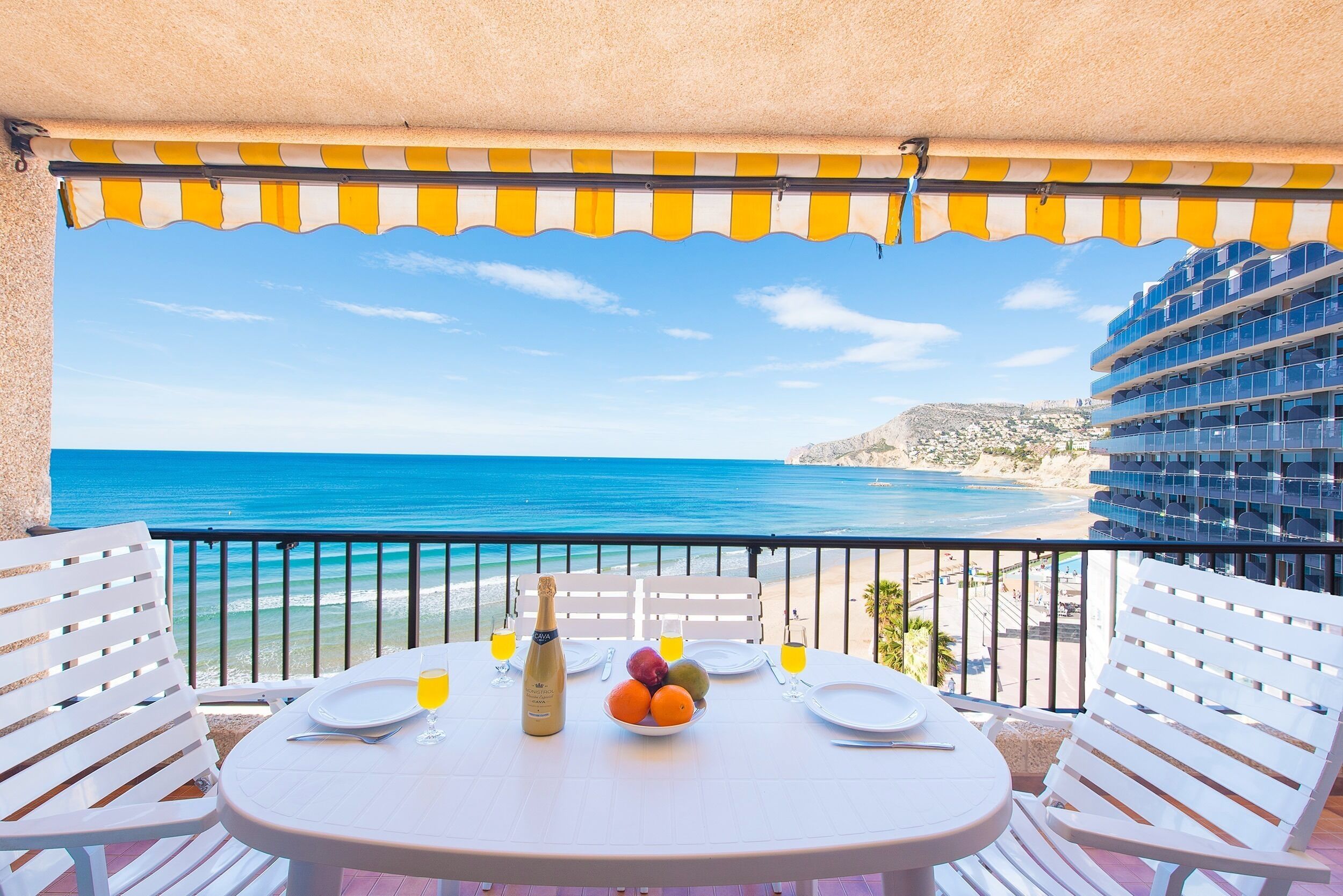 Un balcon avec une table dressée pour deux personnes, donnant sur une plage, un ciel bleu azur et l’océan.