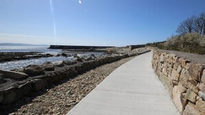 Een betonnen pad langs een rotsachtige kustlijn, met een stenen muur aan één kant en uitzicht op de zee aan de andere kant.