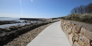 Un chemin de béton longe une côte rocheuse, avec un mur de pierres d’un côté et une vue sur la mer de l’autre.