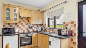 A compact kitchen with wooden cabinets, a built-in oven, a dishwasher, and a window overlooking a garden. - Spiddal Cottage splendid base to tour the west (Spiddal)