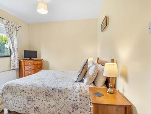 Een slaapkamer met een bed, nachtkastje met een lamp, een dressoir en een televisie op een meubel.