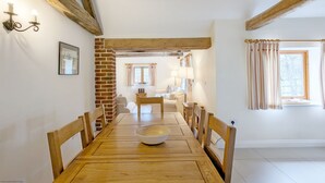 Un coin repas avec une table en bois, des chaises et un saladier. Il y a un mur en briques et une fenêtre avec des rideaux.