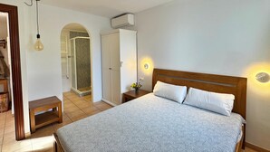 Een slaapkamer met een groot bed, een houten hoofdbord, een nachtkastje, een wandlamp en een douchegedeelte met betegelde wanden.