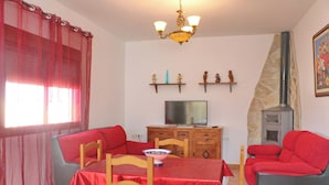 Un salon avec un ensemble de canapés rouges, une table à manger avec des chaises, un téléviseur posé sur un meuble en bois, une cheminée en pierre et un lustre.