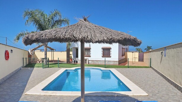 Une piscine surmontée d’un parasol en chaume, entourée de chaises longues bleues et avec un bâtiment blanc en arrière-plan.