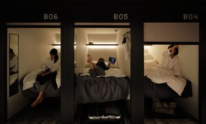 Une chambre d’hôtel avec trois lits, portant les numéros B06, B05 et B04. Chaque lit est équipé d’une table de chevet et d’une lampe.