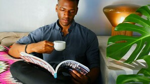 A person sitting on a sofa with a book and a mug, a lamp, and a plant in the foreground.
