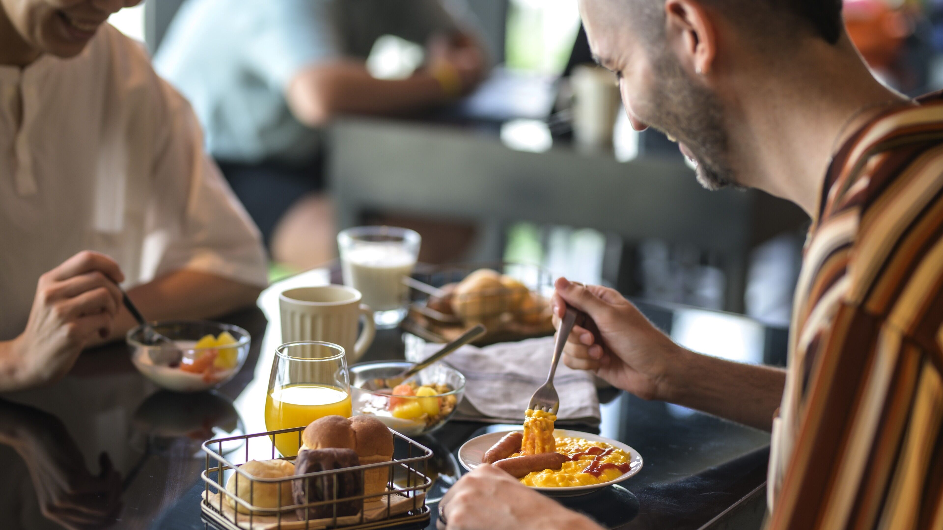 A breakfast setting with a table set for three people, including a basket of pastries, a jug of orange juice, and plates of food.